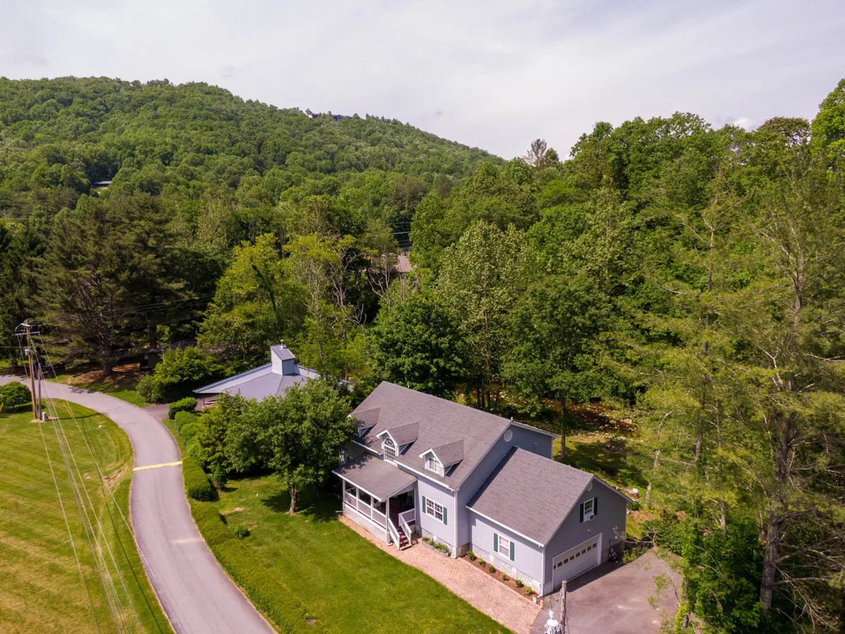 A wide exterior perspective of the home nestled among mature trees, with a visible driveway and welcoming porch. The setting suggests easy access to nature and nearby trails for exploration.