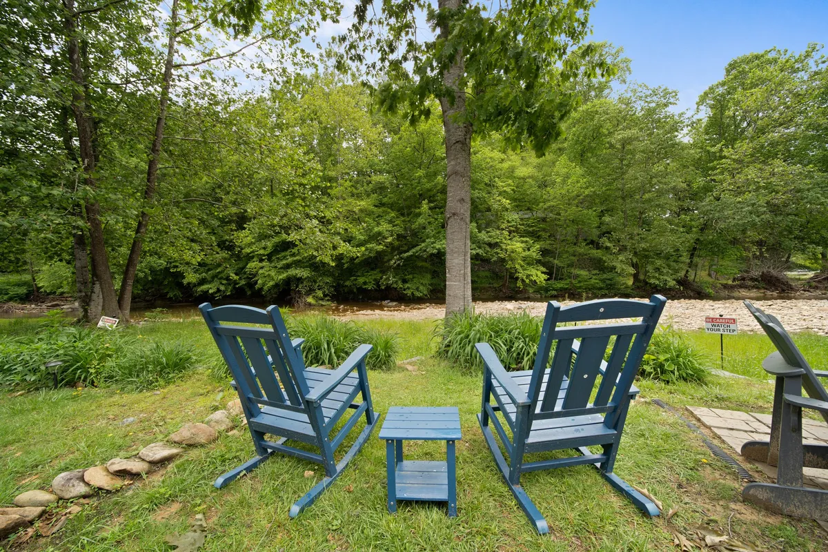 Two inviting blue rocking chairs sit on a porch or deck, positioned beside a small side table. Lush green surroundings and a tranquil river view are visible beyond the chairs, evoking a peaceful, nature-connected atmosphere ideal for relaxing or enjoying the outdoors.