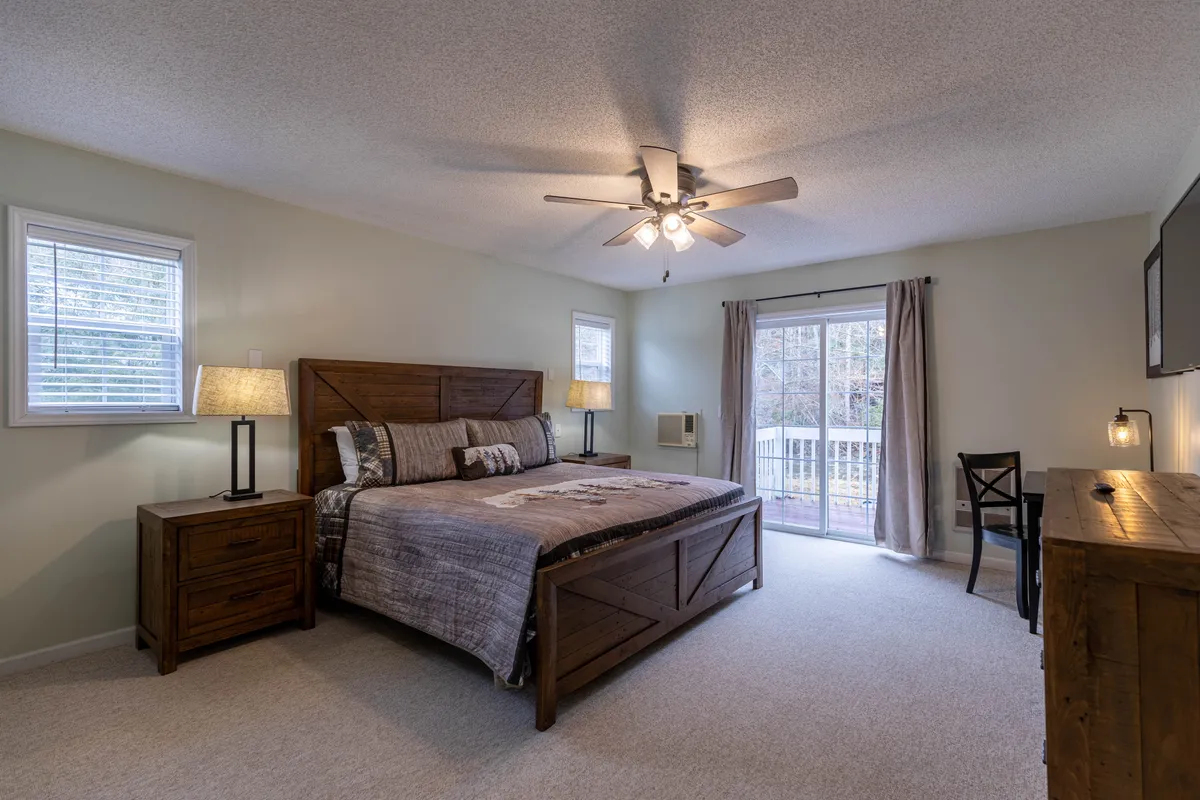 A serene bedroom with a comfortable bed between nightstands, large windows, and a sliding door that bathes the room in natural light. The ambiance is calm and cozy, ideal for relaxing or enjoying peaceful mornings.