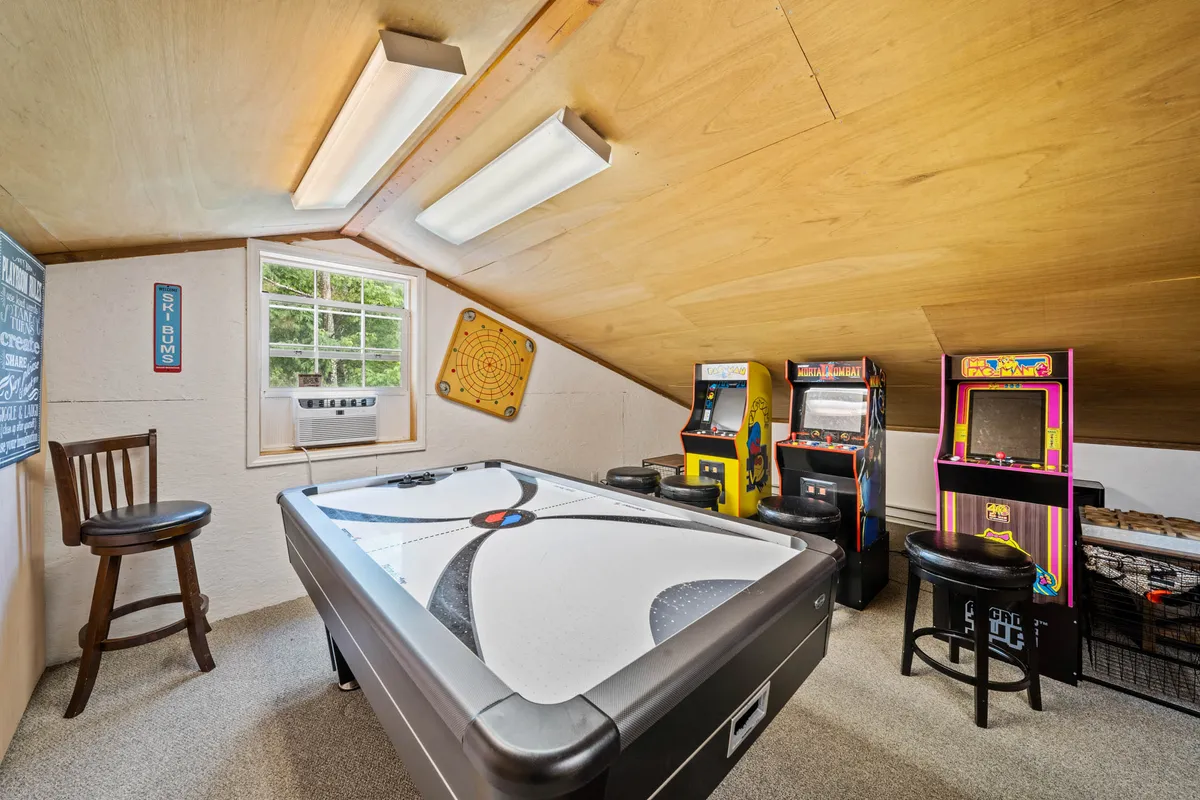 A lively game loft interior featuring an air hockey table, several arcade game machines (including a visible Pac-Man), a dartboard, and a window letting in natural light. The setup invites playful competition and relaxed socializing.