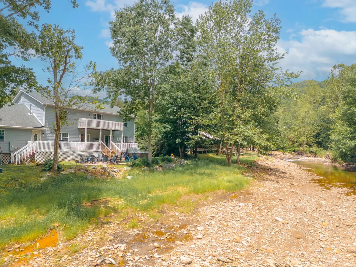 This exterior shot captures the house with a balcony overlooking the riverbank, surrounded by lush greenery. The image hints at direct river access, places for fishing or picnicking, and the serene, natural setting perfect for group relaxation and shared outdoor experiences.