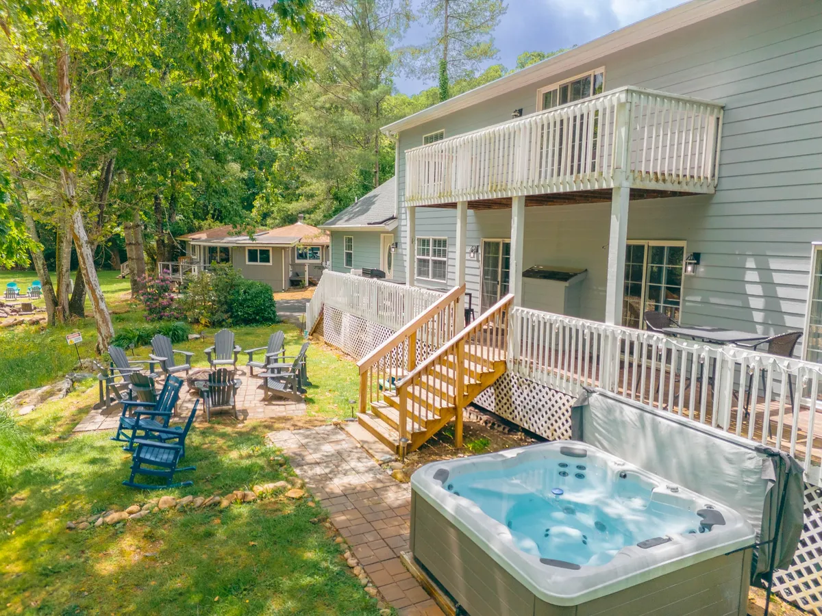 The hero image displays the home's spacious deck nestled among lush trees, featuring a bubbling hot tub, comfortable outdoor seating, and a firepit area perfect for gatherings. The Watauga River is nearby, and the scene captures the warm glow of sunset, inviting relaxation and outdoor fun.