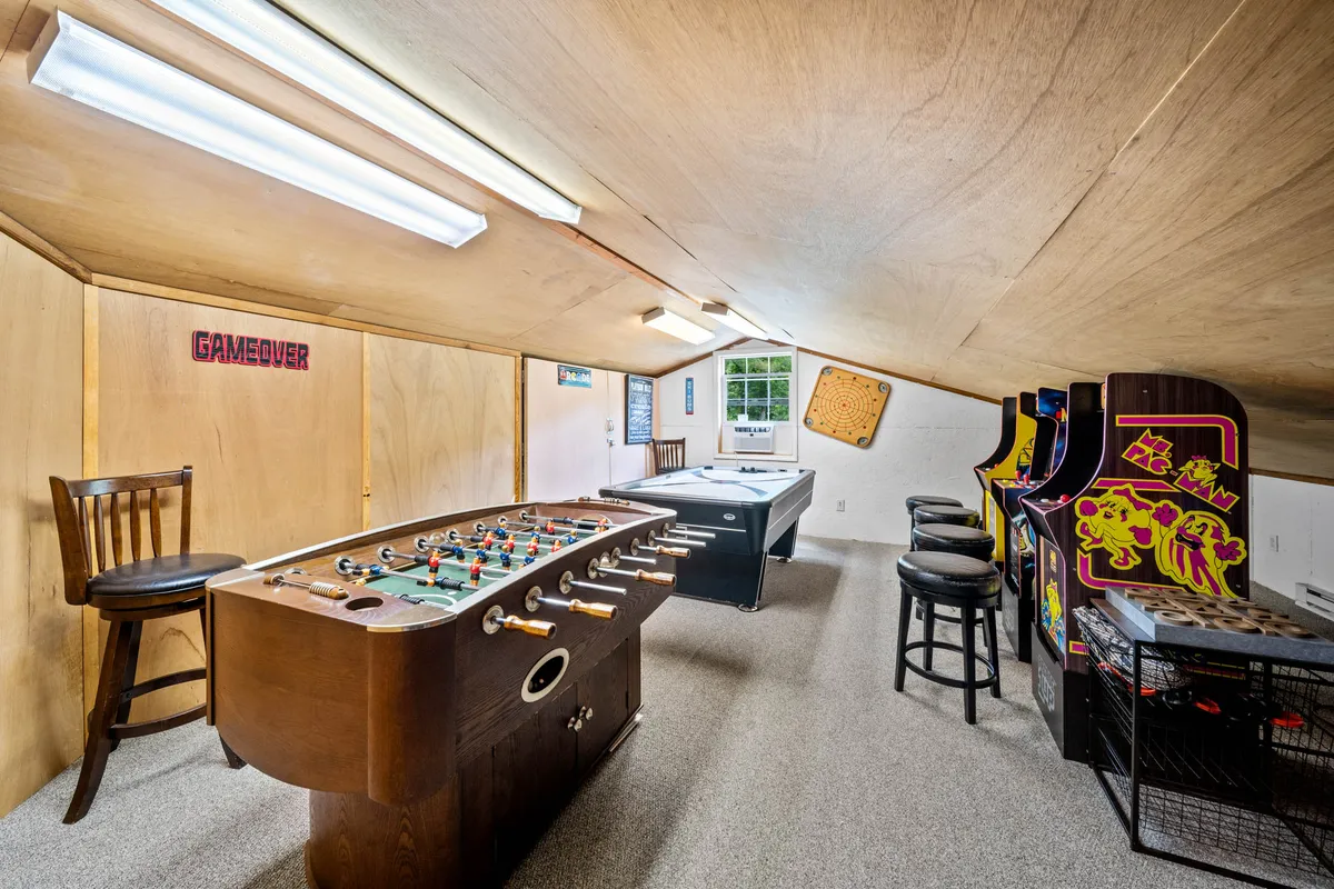 A vibrant game room featuring a foosball table, pool table, and several arcade machines including classic games. Bar stools line one side, creating a comfortable space for socializing. The room exudes a fun and energetic vibe, ideal for family gatherings and friendly competition.