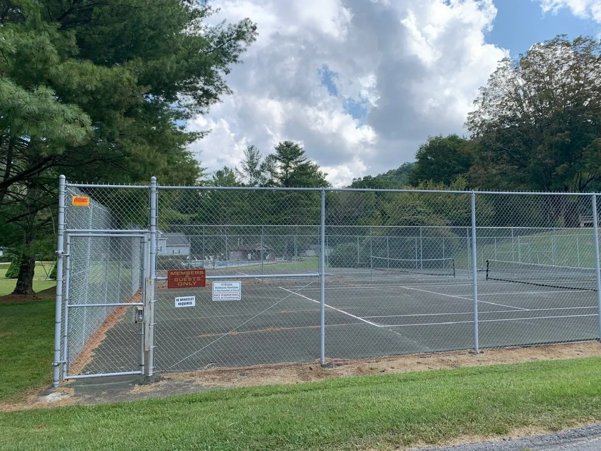 A tennis court surrounded by a chain-link fence and bordered by leafy green trees. The scene is active and communal, inviting guests to engage in outdoor sports or enjoy the vibrant neighborhood atmosphere.