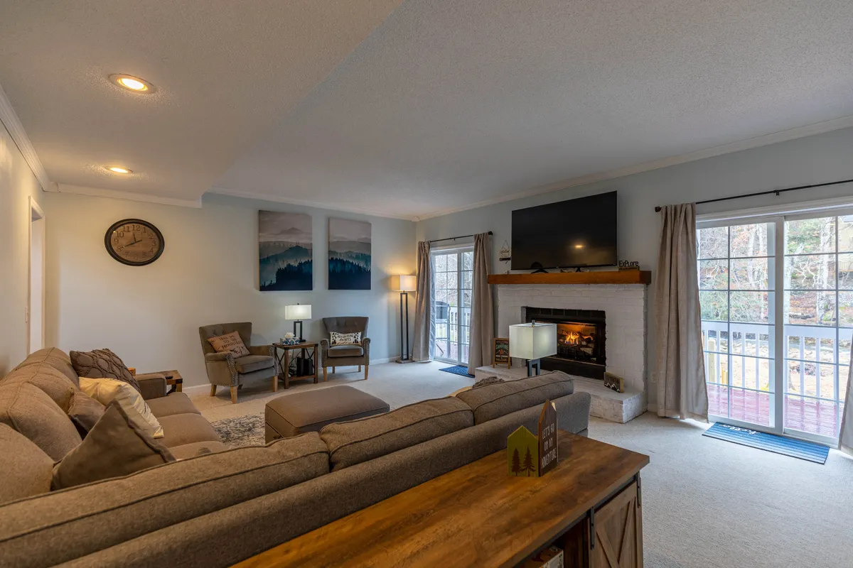 A spacious, warmly lit living room with a large sectional sofa arranged around a fireplace, a coffee table at the center, artwork on the wall, and expansive windows letting in natural light. The atmosphere is inviting, perfect for relaxing, socializing, or watching TV after a day outdoors.